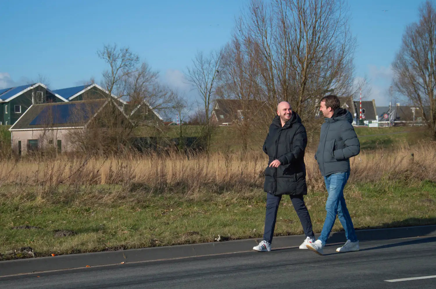Twee mannen die naast elkaar lopen op een geplaveide weg, de een in een zwarte gewatteerde jas en witte sneakers, de ander in een donkergrijze capuchonjas en blauwe jeans.