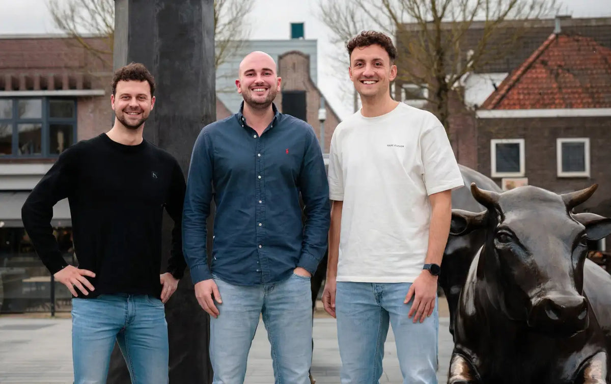 Drie mannen staan samen buiten, gekleed in casual kleding, waaronder een zwart shirt met lange mouwen, een marineblauw overhemd met rode borduursels op de borstzak en een wit t-shirt.