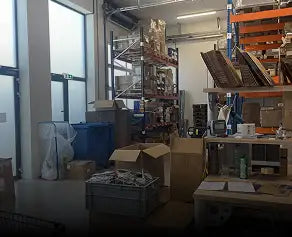De afbeelding toont een rommelige magazijnruimte met planken, dozen en apparatuur.