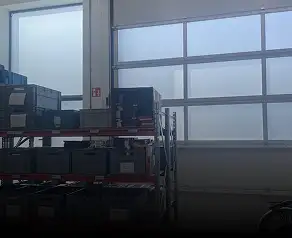 De afbeelding toont een magazijninterieur met metalen stellingen waarop gestapelde zwarte dozen staan, opgesteld voor grote matglazen ramen.