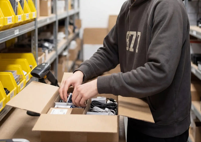 Een persoon in een donkergrijze hoodie met beige „bzi”-opschrift is elektronische componenten aan het hanteren in een open kartonnen doos.