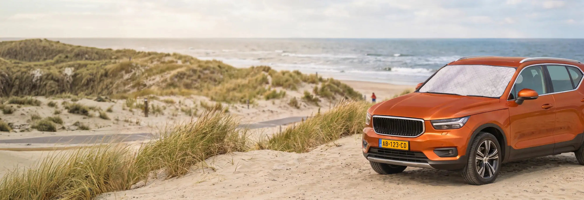Een oranje Volvo XC40 SUV geparkeerd op zanderige duinen bij de oceaan.