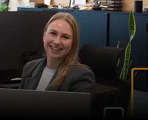 Een glimlachende vrouw met blond haar die een grijze blazer over een witte top draagt, zittend achter een bureau op kantoor.