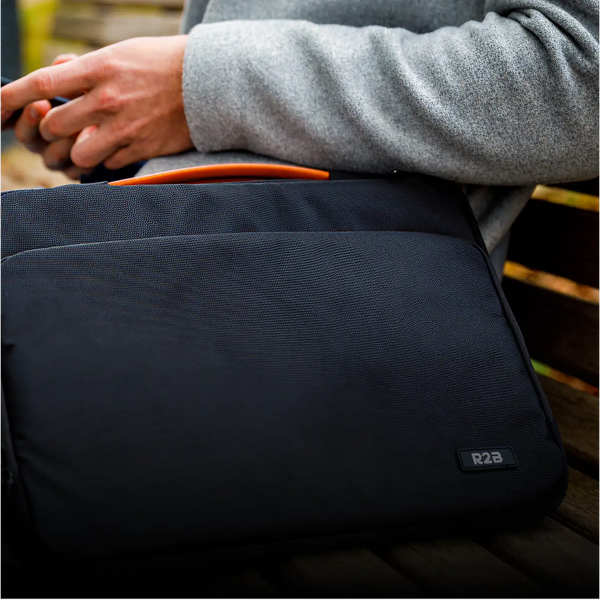 Zwarte, gestructureerde laptoptas met oranje handgreep en een klein r2b-logo patchje.